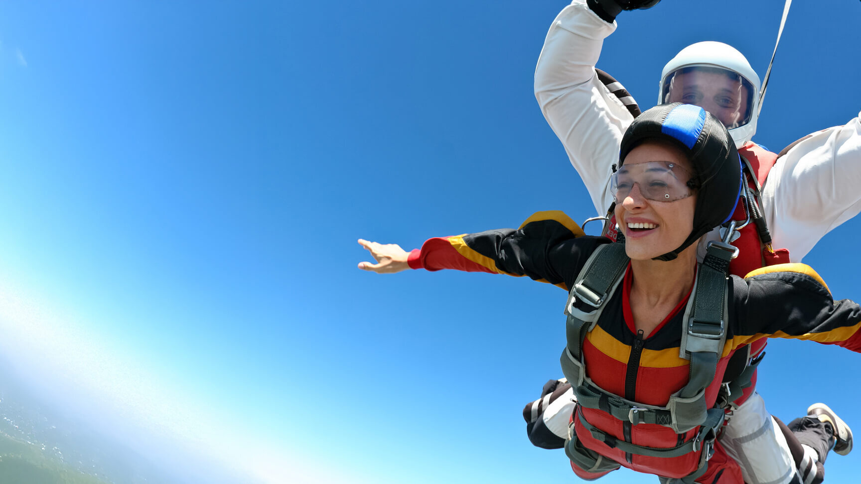 Smiling woman tandem skydiving with an instructor, arms outstretched in freefall under a bright blue sky.