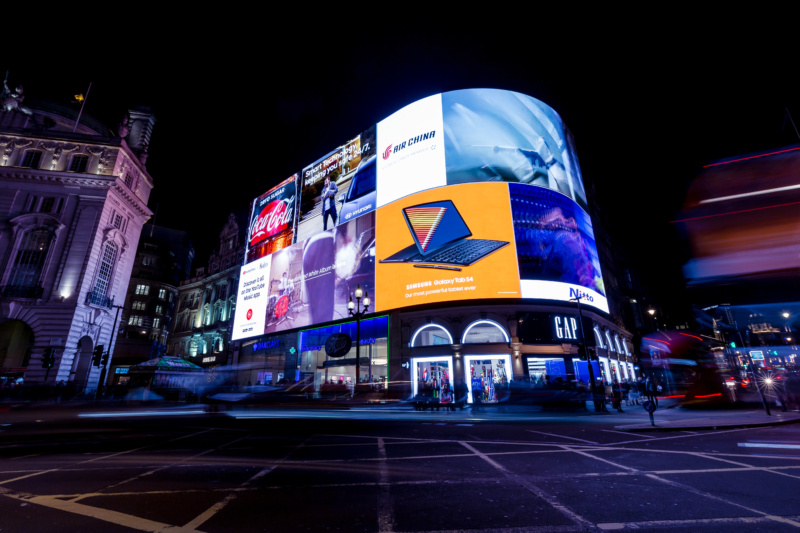 Illuminated digital billboards at night, featuring advertisements for Coca-Cola, Air China, Samsung Galaxy Tab S4, Hyundai, and other brands, with light trails from passing traffic and people walking below.