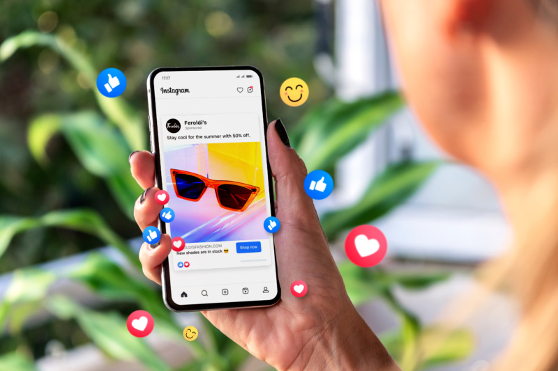 Person holding a smartphone showing an Instagram ad for red sunglasses with 50% off, surrounded by floating social media reaction icons like hearts, thumbs up, and smiley faces.