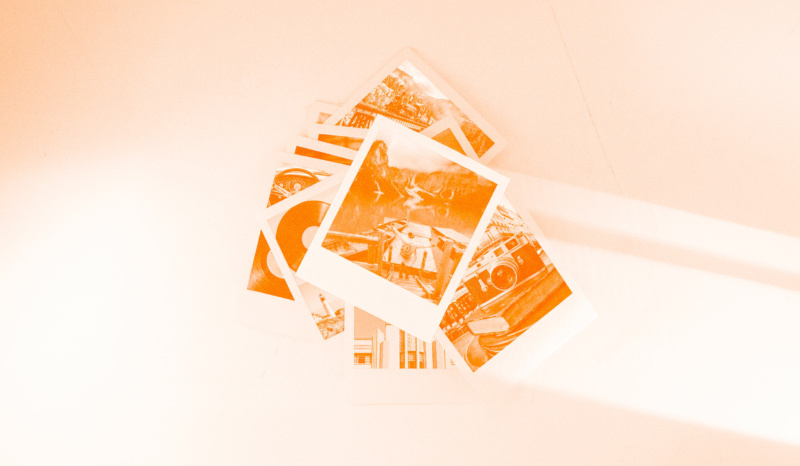 Stack of orange-tinted printed photos on a light surface, featuring travel and lifestyle scenes like landscapes, a boat, and a vintage camera.