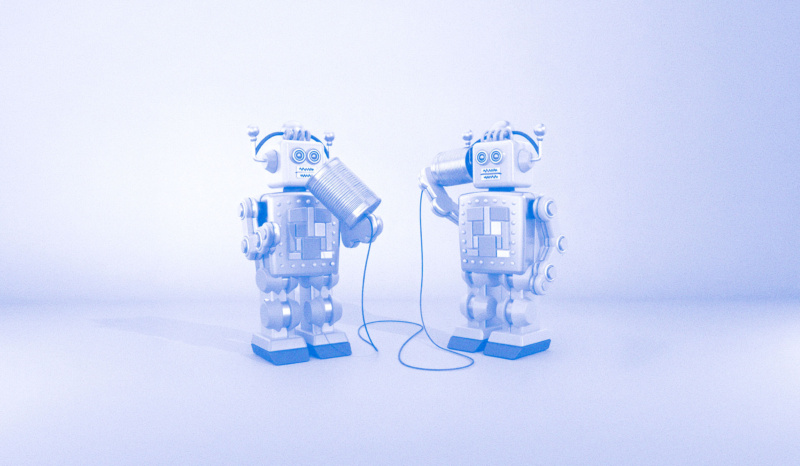Two toy-style robots face each other on a pale background, communicating with a tin-can telephone connected by a string.