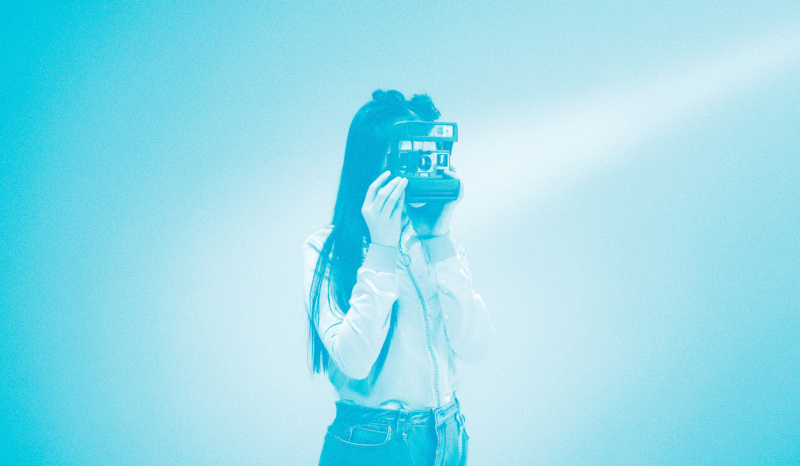 Teal-toned image of a person holding a vintage instant camera up to their face, capturing a moment with analog technology.