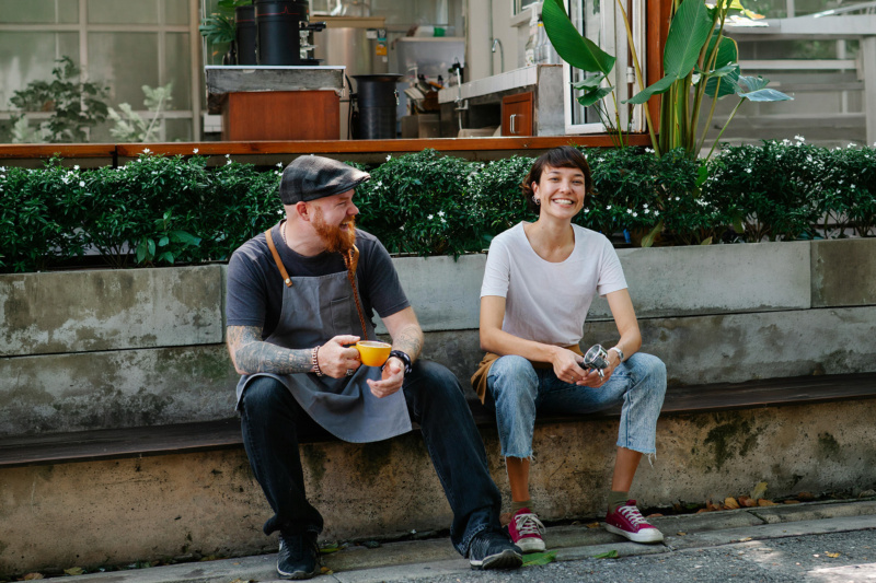 Two café workers sitting outside on a bench, smiling and chatting while holding coffee tools in a leafy patio setting.