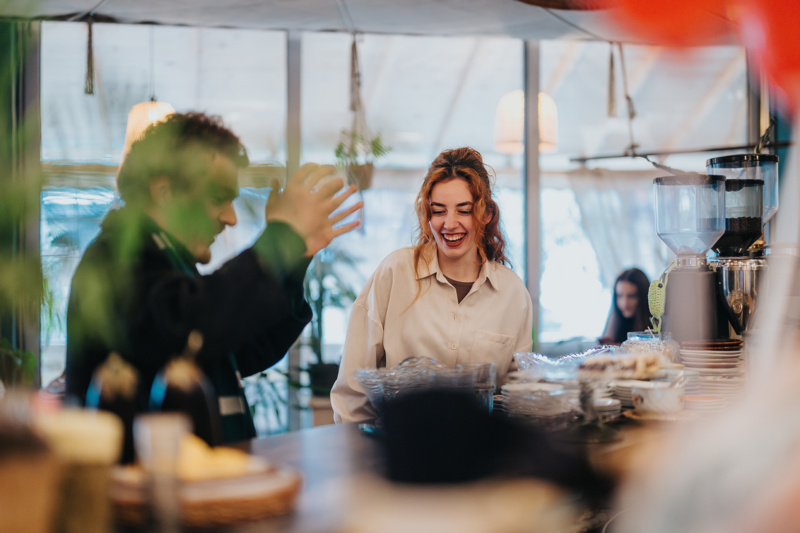 Barista laughing with a customer across a café counter, with coffee equipment and pastries in a warm, inviting interior.