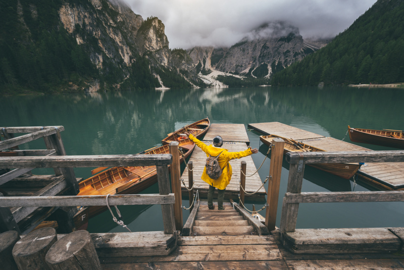Person in a yellow jacket standing on a wooden dock with arms outstretched, facing a calm mountain lake surrounded by forest and misty peaks.
