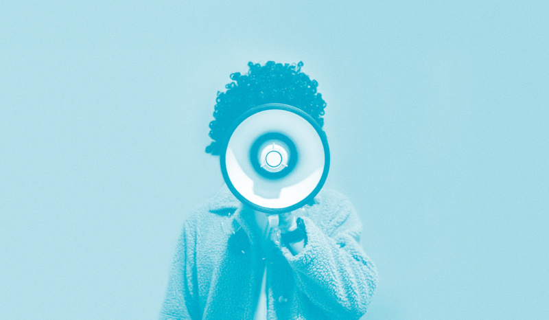Person with curly hair holding a megaphone directly in front of their face against a plain blue background, obscuring their features.