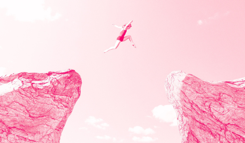 Person mid-air leaping across a wide gap between two rocky cliffs against a cloudy sky, with the image tinted pink.