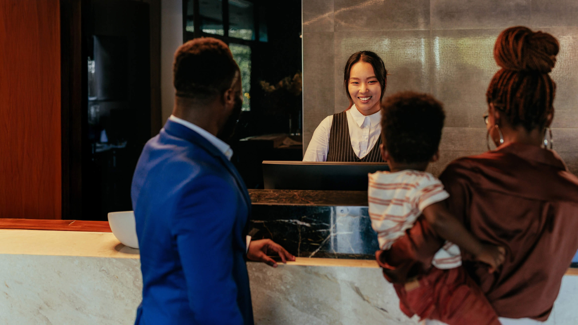 Hotel front-desk scene with a smiling receptionist greeting a family. A man in a blue suit stands at the counter while a woman holding a young child faces the desk, creating a warm, welcoming check-in moment.