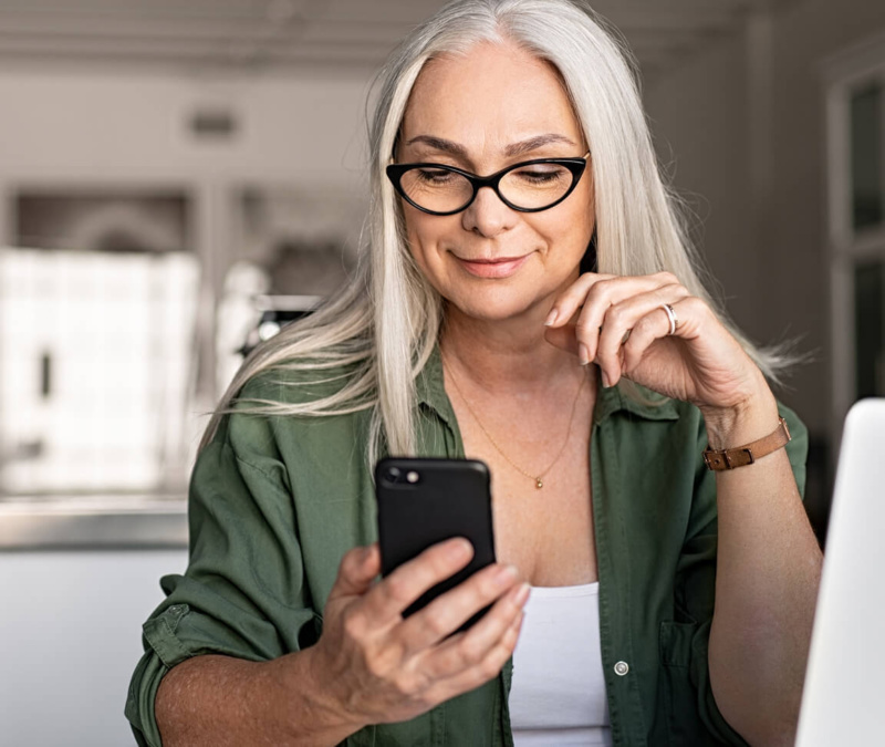 Older woman with long grey hair and glasses smiles while checking her smartphone at home.