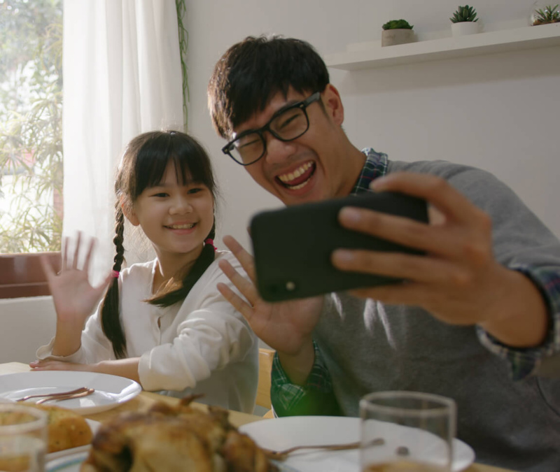Father and daughter smiling and waving while taking a selfie at the dinner table in a sunlit home.