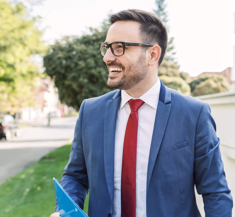 Smiling REALTOR® in a blue suit and red tie walking outdoors, holding a blue folder, with greenery and houses in the background.