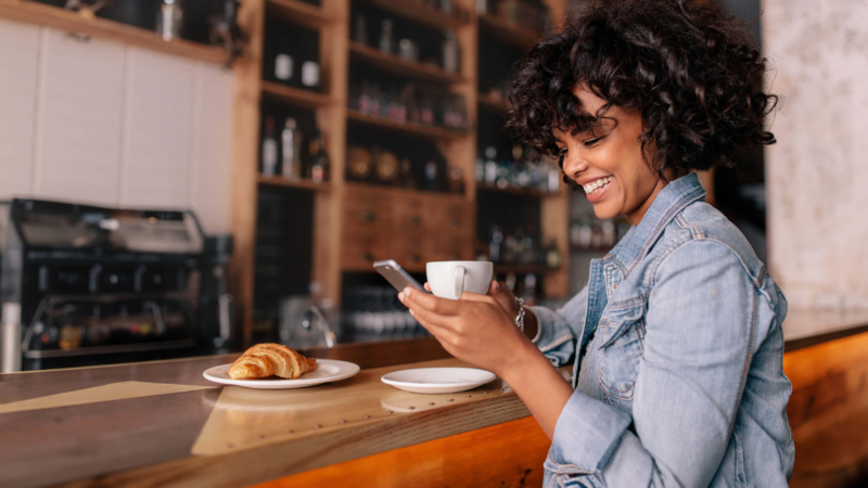 Woman sitting at a café bar, smiling at her phone while holding a coffee, with a croissant on a plate beside her.