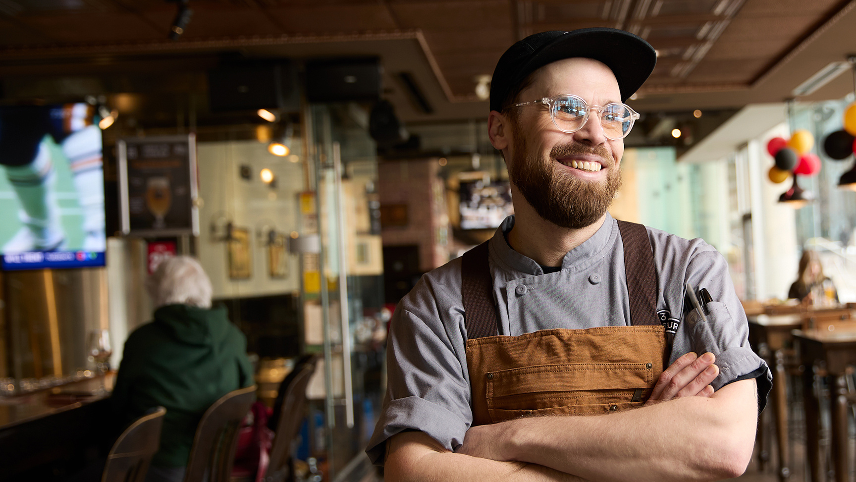 Smiling chef with arms crossed stands inside a modern restaurant, wearing glasses, a cap, and a brown apron.