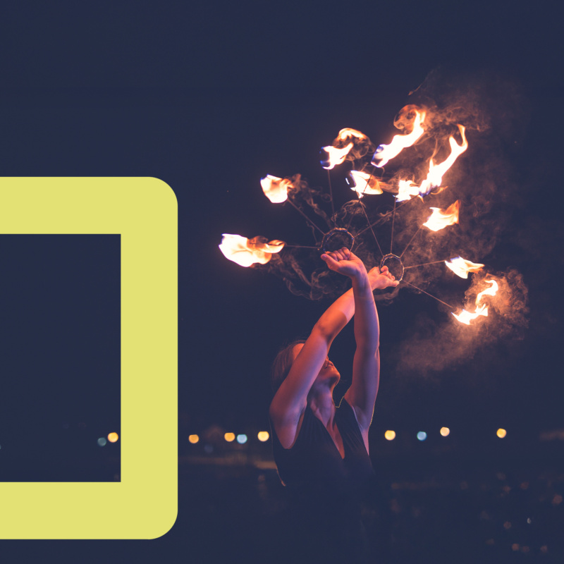 Performer holding a circular fire prop with flames against a dark background, partially framed by a yellow corner shape.