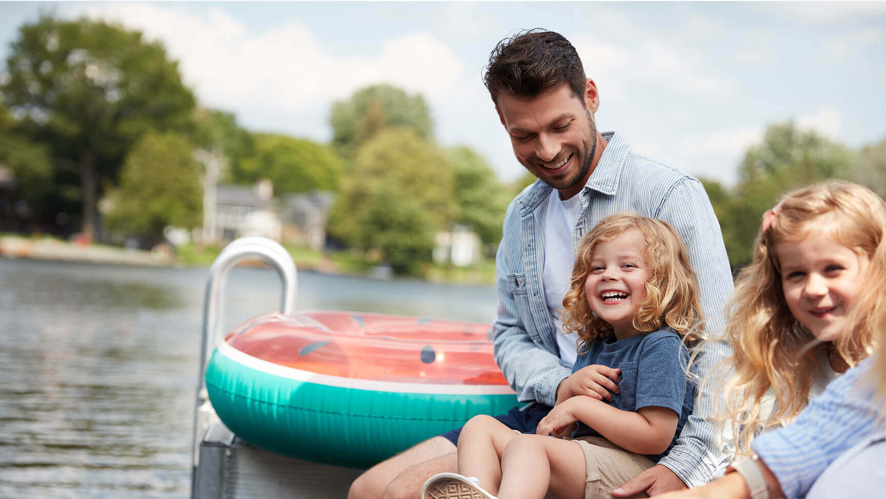 Father and two young children sitting on a dock by the water, smiling and enjoying a sunny day outdoors.