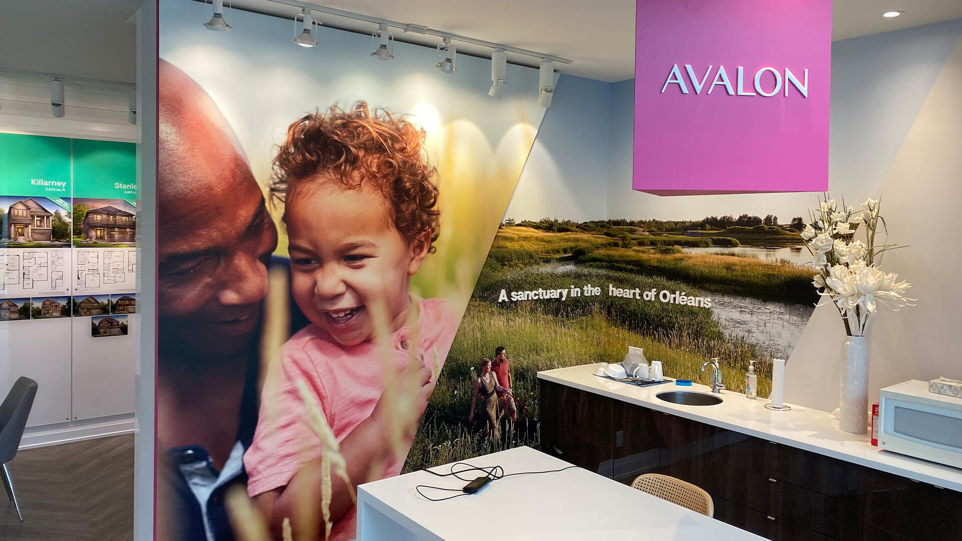 Interior of the Minto Avalon sales centre showcasing a feature wall with large lifestyle imagery and the tagline “A sanctuary in the heart of Orléans.” A bright pink pendant light, adorned with the Avalon logo, hangs from the ceiling.