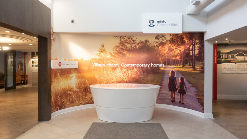 Interior of the Minto Mahogany sales centre featuring a display wall with the tagline “Village charm. Contemporary homes.” and a mural of two children walking along a path at sunset.