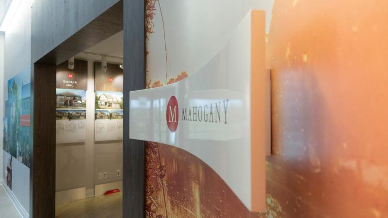 Side view of the Mahogany logo sign extending from a wall mural depicting a warm lakeside scene inside a Minto Communities sales centre.