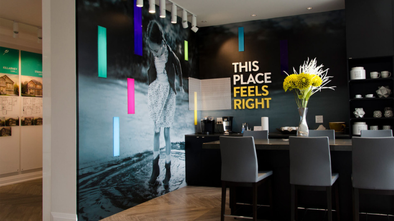 Wall mural inside the Minto Quinns Point sales centre featuring a black-and-white photo of a child playing in water with colourful accent lighting and the tagline “This place feels right.”