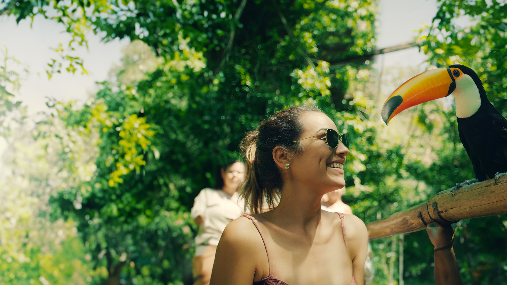 A woman wearing sunglasses smiles at a toucan perched on a wooden railing in a lush, sunlit rainforest setting.