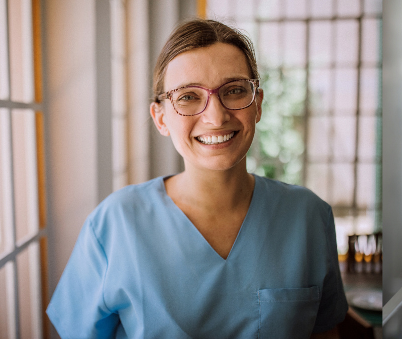 Smiling healthcare worker in blue scrubs wearing glasses, standing indoors by windows.
