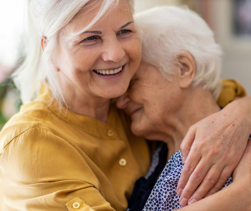 Older woman in mustard shirt hugging another elderly woman with a smile.