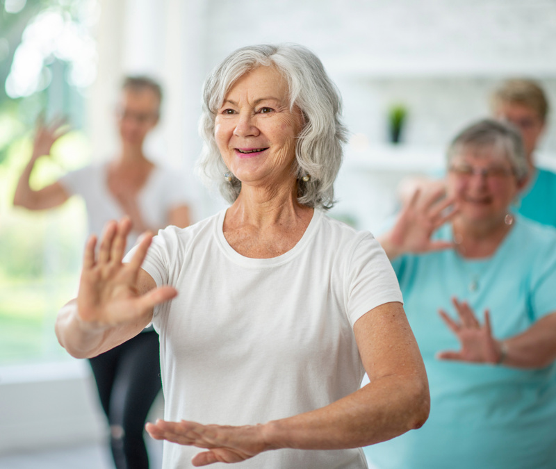 Older woman in white shirt doing tai chi with a group of seniors in background.