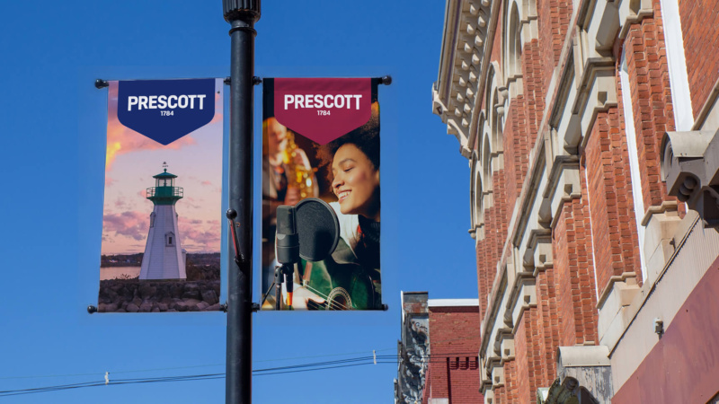 Street banners on a lamppost featuring Prescott branding—one with a lighthouse at sunset, the other with a musician singing—against a clear blue sky and historic brick building.