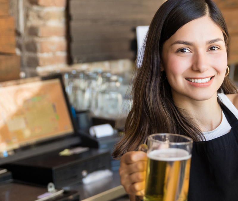 Smiling bartender holding a glass of beer in a warmly lit bar setting, with taps and equipment behind them.