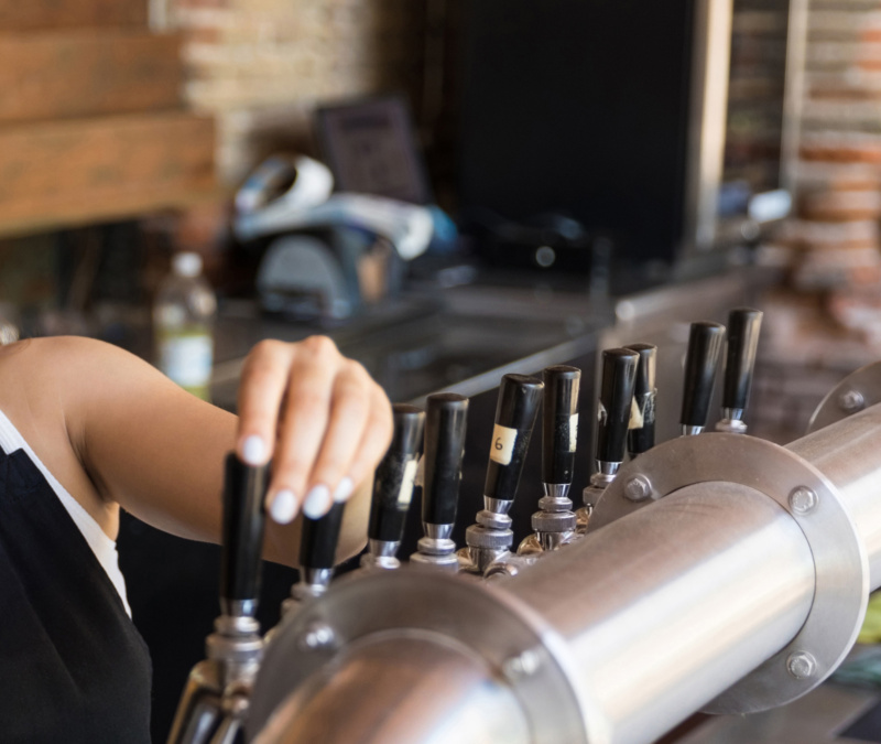 Smiling bartender holding a glass of beer in a warmly lit bar setting, with taps and equipment behind them.