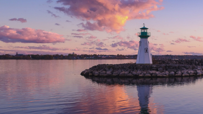 A tranquil sunset scene at the Prescott lighthouse, perched along a rugged rocky shoreline, where soft pink clouds drift across the sky and mirror perfectly in the glassy, still water.