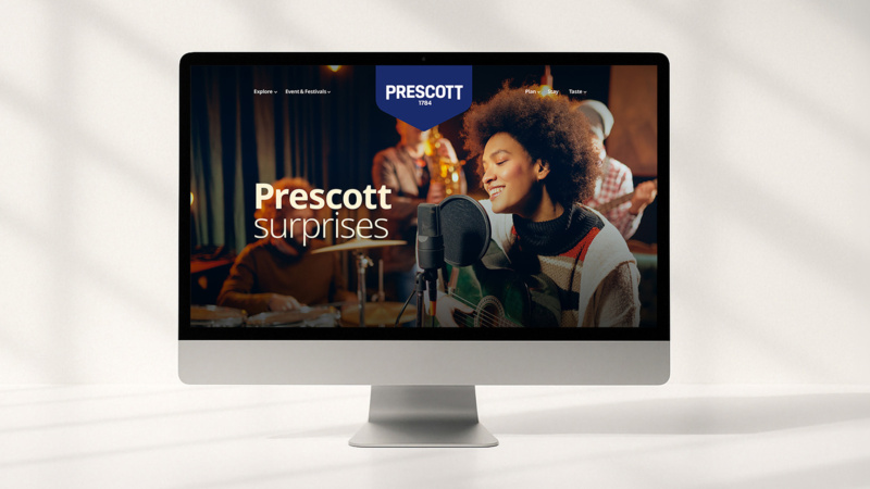 Desktop computer displaying the Prescott website homepage featuring a musician and the text “Prescott surprises”.