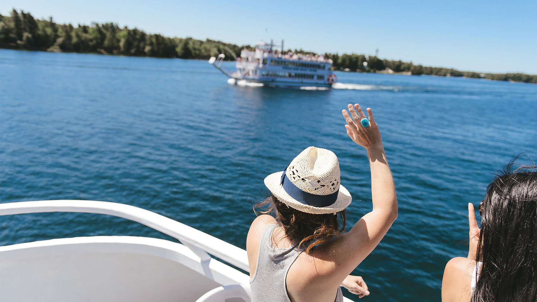 Two people on a boat wave to a passing cruise ship on a sunny day in the Thousand Islands.