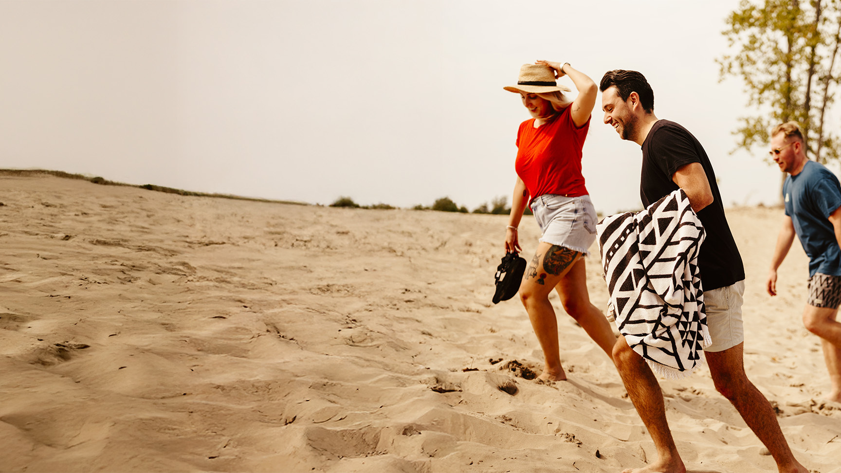 Three friends walking barefoot up a sandy beach hill on a sunny day, carrying towels and beach gear.