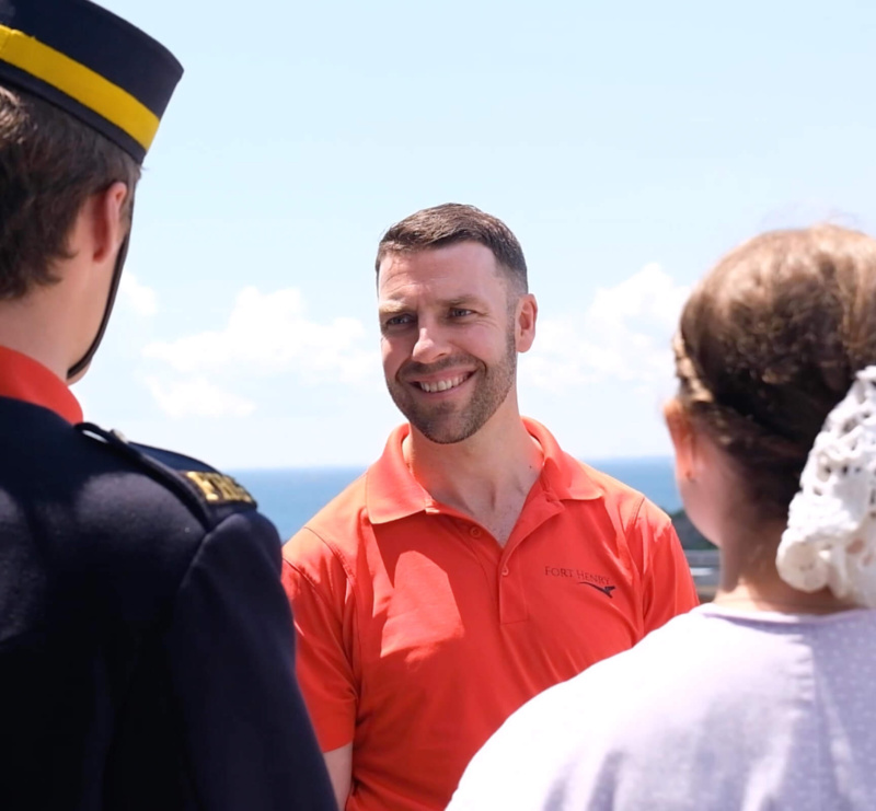 A smiling man in a red Fort Henry polo shirt talks with two people, including a costumed interpreter, outdoors with the blue sky and water in the background.