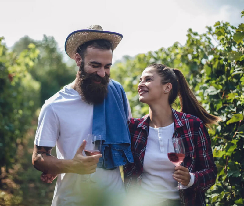 Smiling couple walking through a vineyard, holding wine glasses on a sunny afternoon.