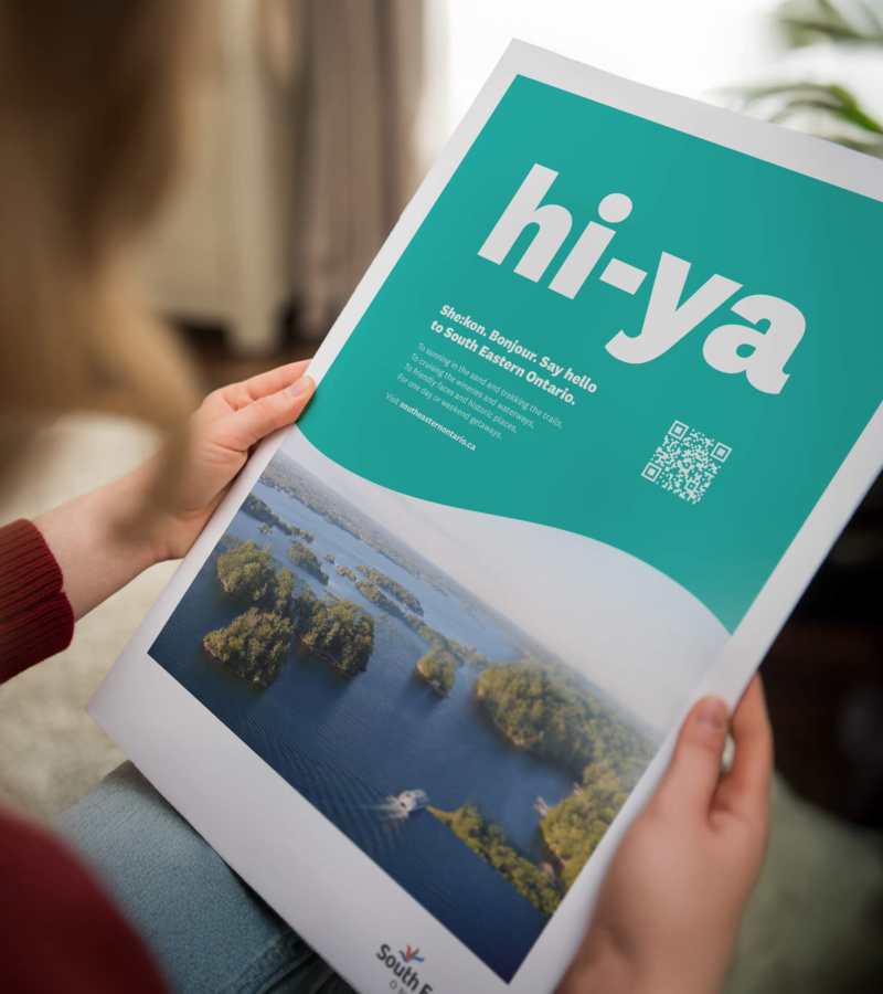 Person holding a print ad with aerial view of islands and text reading “hi-ya” promoting South Eastern Ontario.