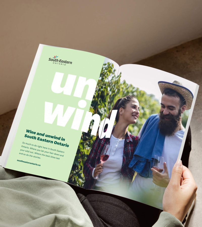 Person reading a South Eastern Ontario magazine ad with the headline “Wine and Unwind” and a photo of a smiling couple enjoying wine in a vineyard.