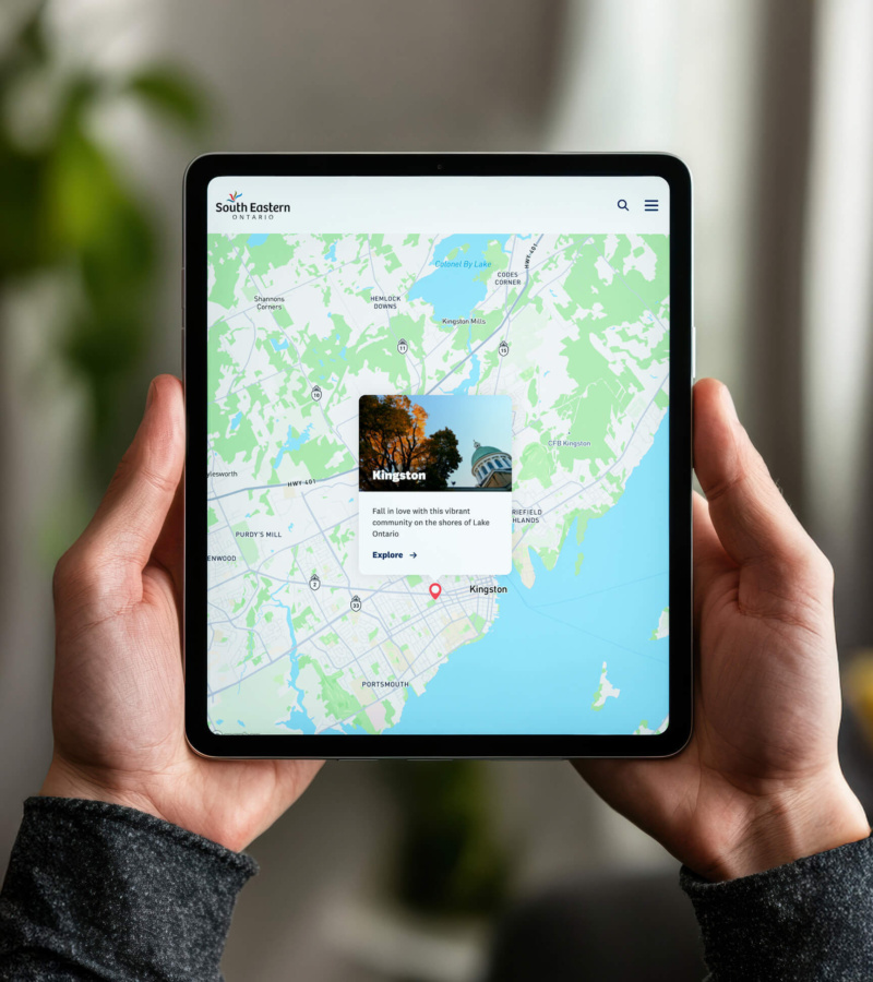A person holds a tablet displaying the South Eastern Ontario website, showing a map of Kingston with a feature card inviting users to explore the city.
