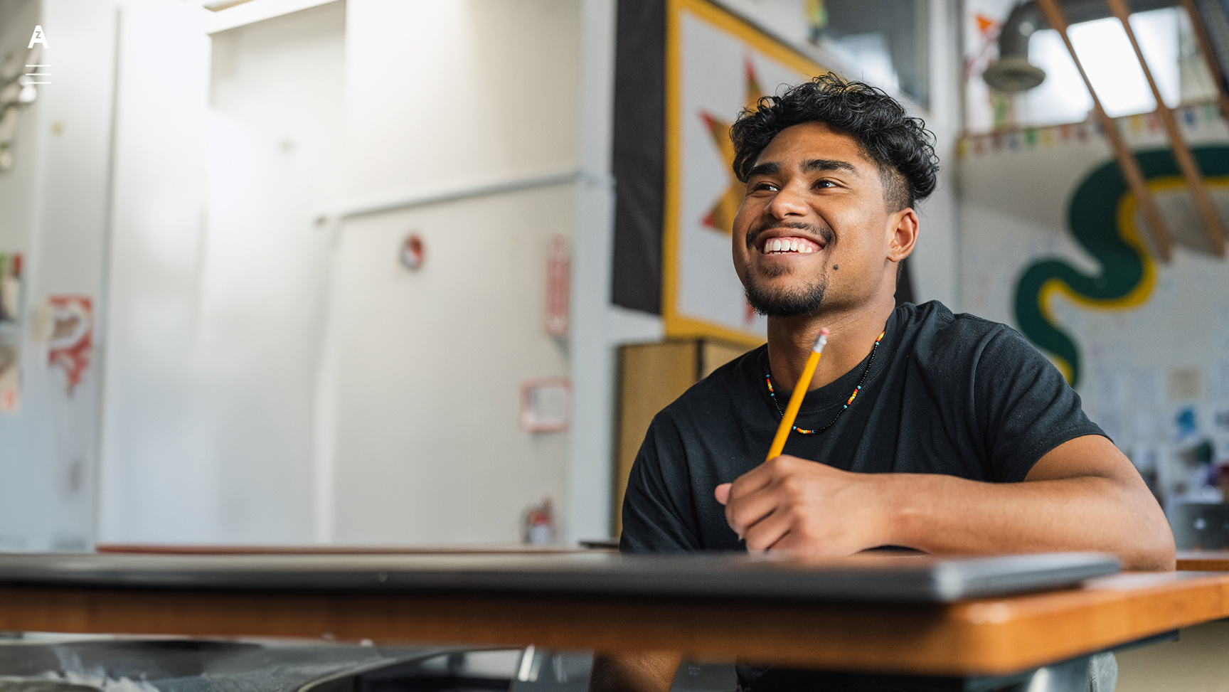 Young man smiling at a desk with pencil in hand in a classroom setting.