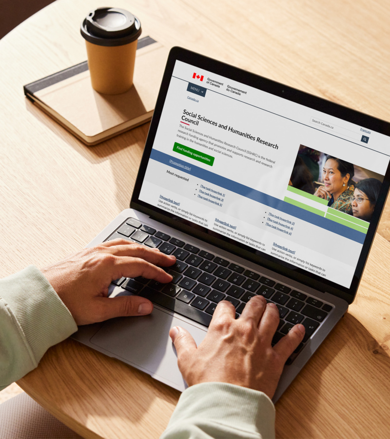 Person using laptop to view SSHRC/CRSH government website at a desk with coffee.