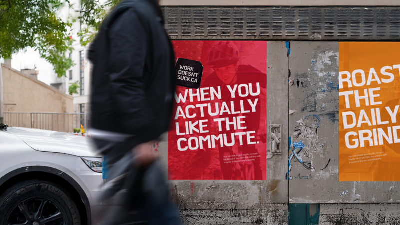 Street posters for WorkDoesntSuck.ca on a worn wall, with bold slogans ‘When you actually like the commute’ and ‘Roast the daily grind,’ as a person walks past.